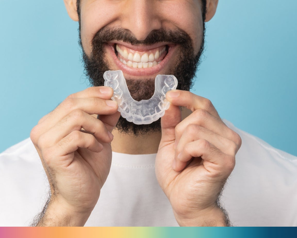 A person with a beard is smiling and holding a retainer in front of their mouth against a blue background.