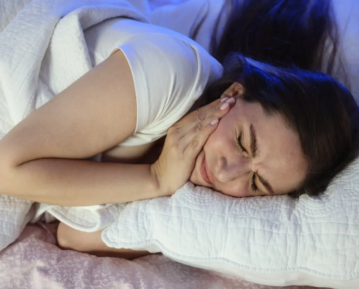 A woman lying in bed at night, holding her jaw in pain and unable to sleep due to severe toothache, highlighting the impact of dental issues on sleep quality.
