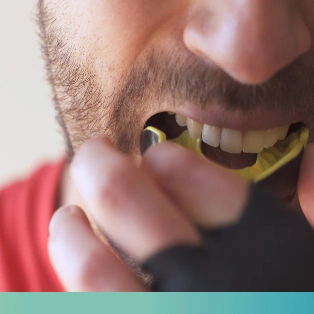 Close-up of a person, their hand partially obscuring their face, placing a yellow mouth guard on their upper front teeth.