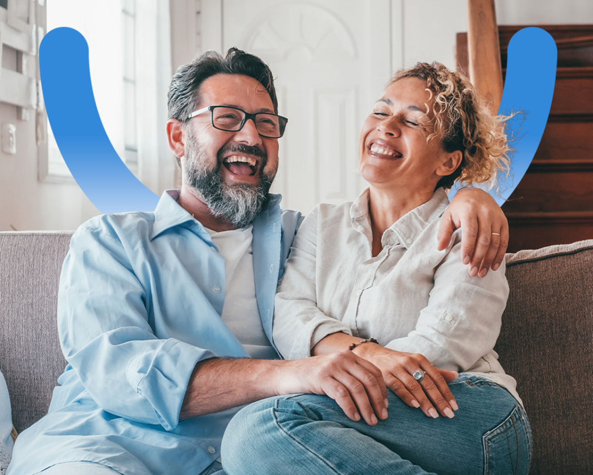 A couple, both wearing light-colored shirts, sit closely on a sofa, smiling and laughing. A blue abstract smile shape is visible behind them.