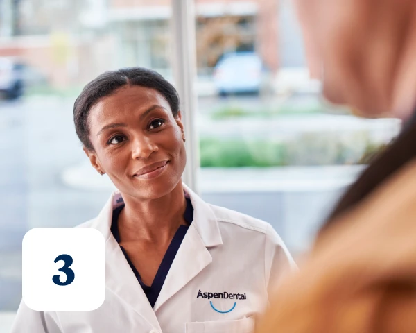 A dental professional wearing an Aspen Dental lab coat converses with a person; the number "3" is visible in the bottom left corner of the image.