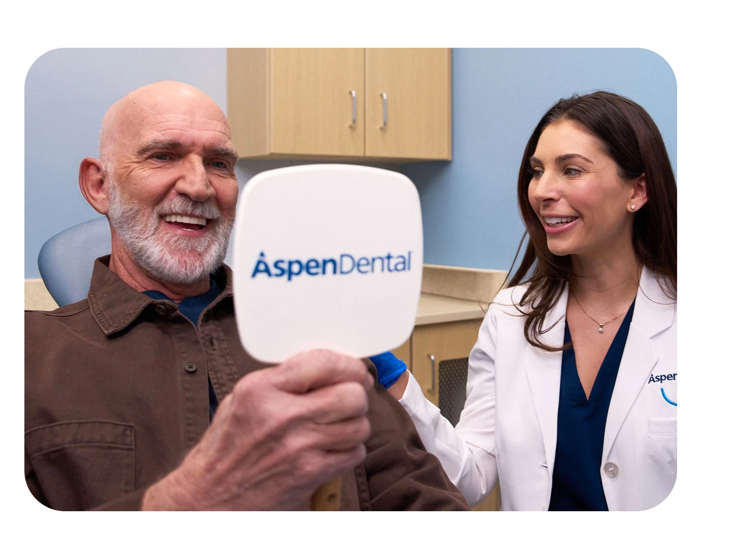 An older man with a bright smile examines his reflection in a handheld mirror while a female dental professional looks on supportively. The image emphasizes how implant dentures can enhance your smile and oral health.