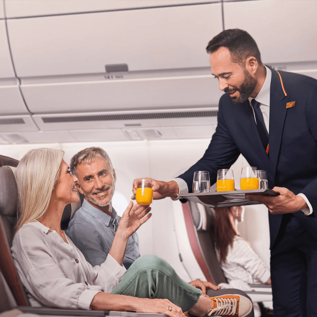 A uniformed Iberia crew member hands two passengers a drink while onboard an aircraft