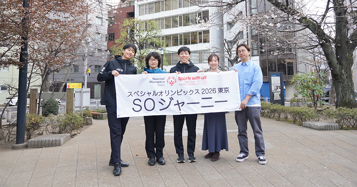 集合写真（後編）：SON様オフィスをバックに桜の咲き始めた公園で、対談メンバー（左から）猪熊氏、仲江氏、三上氏、佐藤、森が笑顔で「スペシャルオリンピックス2026東京」、「SOジャーニー」と書かれた横断幕を持っている様子