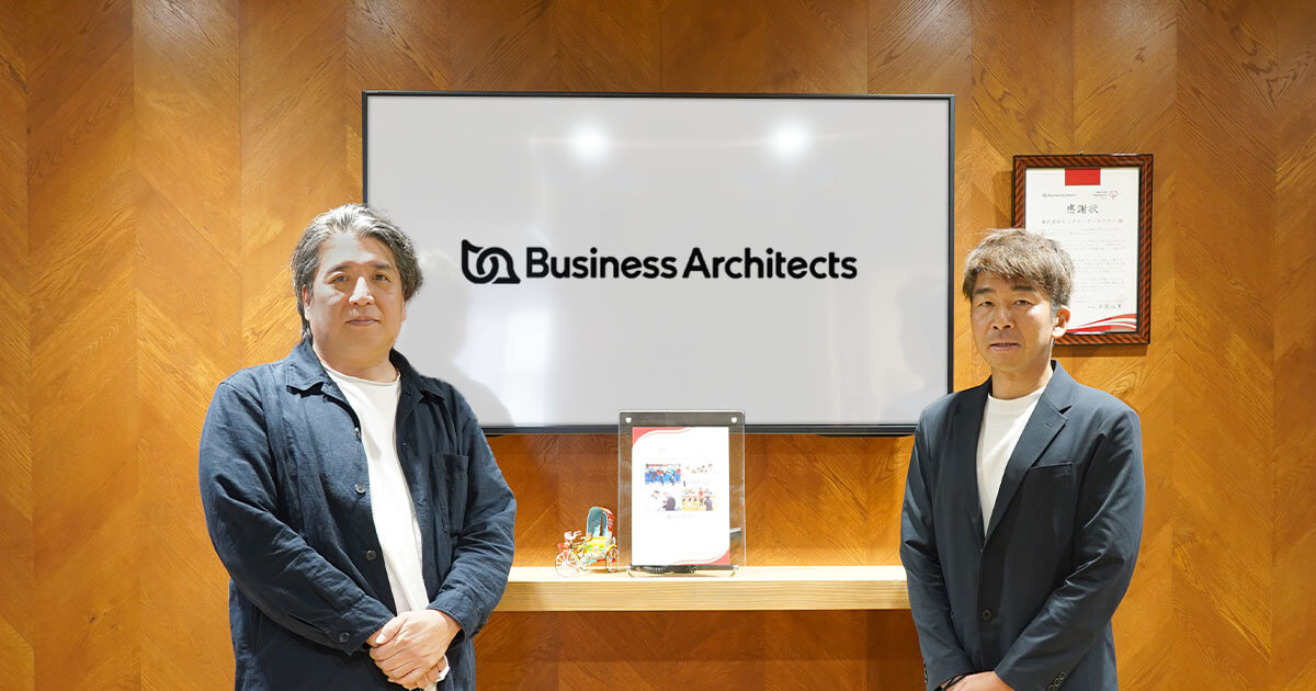 写真：オフィスエントランスをバックにねぎお社長と小山の集合写真