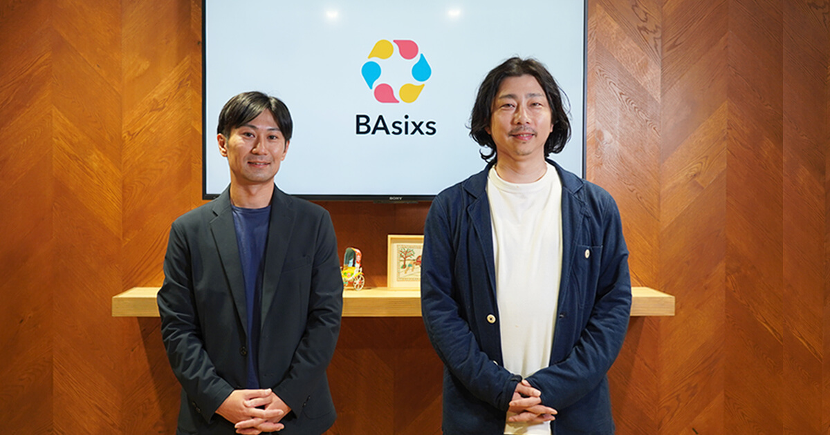 写真：BAsixsロゴのあるエントランスで石田氏と森が並んでいる
