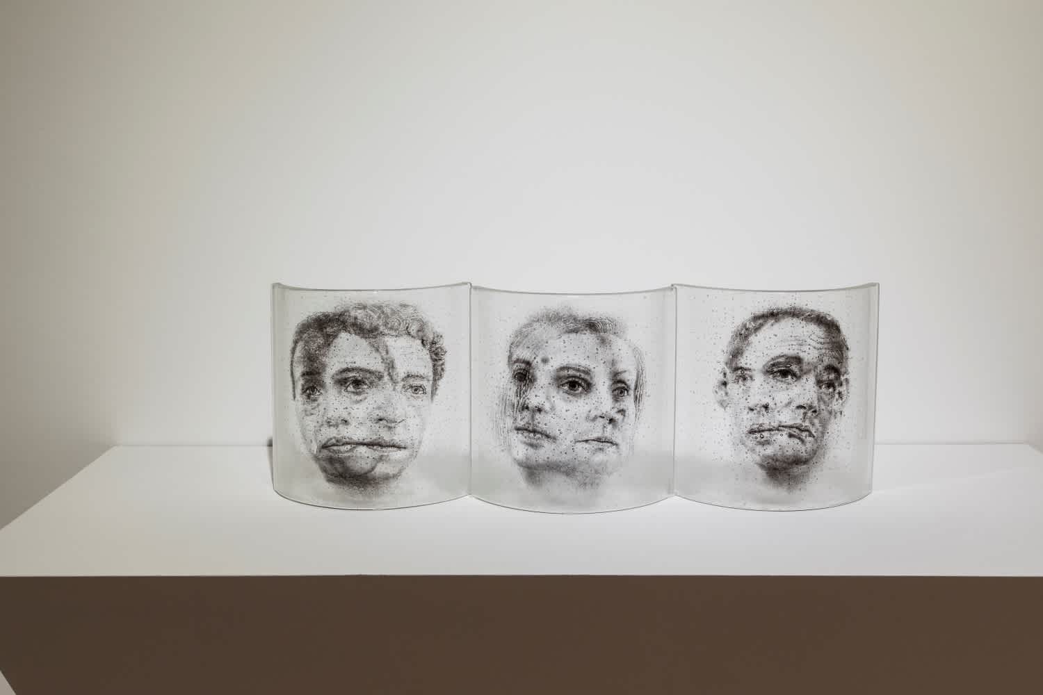Image of Michael Janis, My Other Self: Sanctuary. Three see-through glass objects sit on a white pedestal. The glass holds black images of mirror-like portraits. 