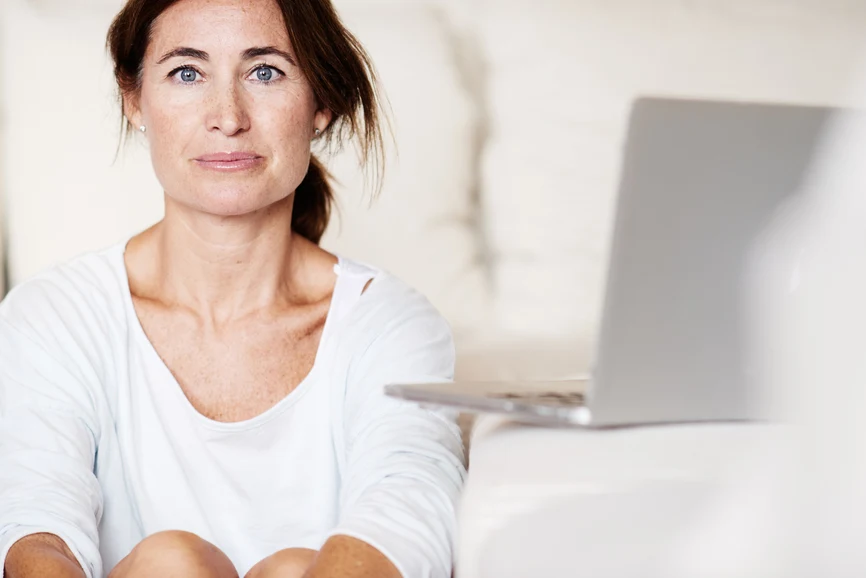 Blue eyed brunette middle-age woman sitting on the floor looking at camera  with neutral expression next to open laptop. AW271 