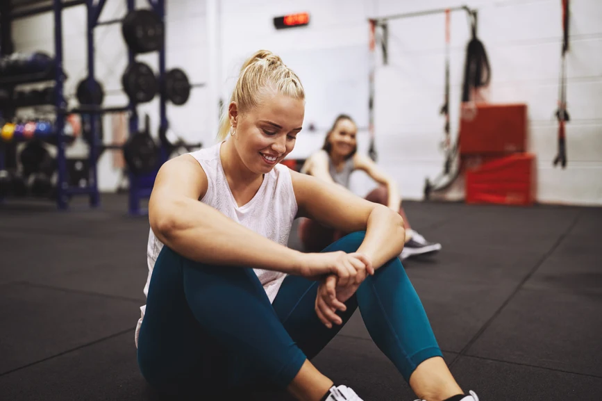 Woman smiling and resting while seated after exercising in gym with a friend. AW149 
