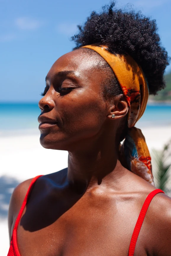 Woman of color in profile on a tropical beach, eyes closed in swimsuit wearing hair wrap. AW156 