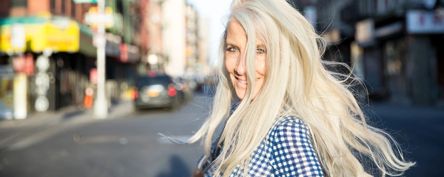 Woman with long white hair looking back smiling on city street, smiling with sun in her face.