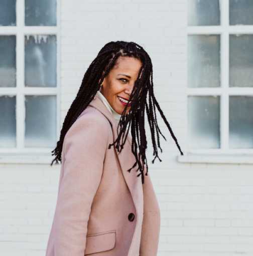 Woman with dreadlocks and blazer, smiling while tossing her hair.