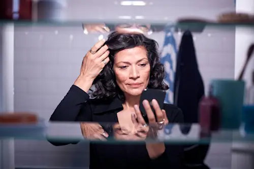 Mature Latina woman checking her hair in her phone while standing in front of bathroom mirror. AW235