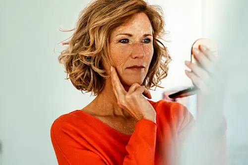 Middle-aged woman in orange top scrutinizing her skin in a compact mirror. thumbnail