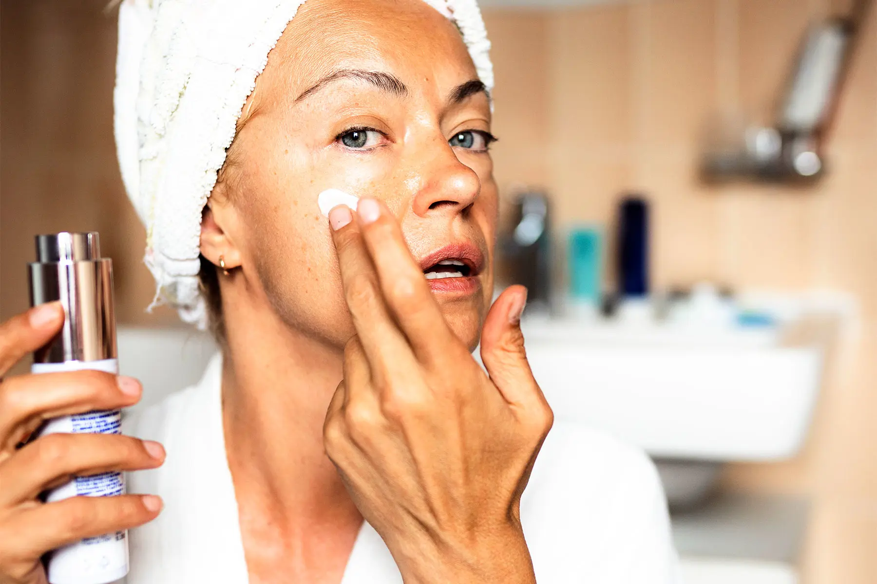 Blue-eyed woman with towel turban and bathrobe holding skin cream in one hand, while applying cream to her face. AW557