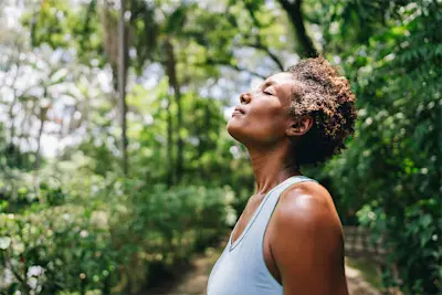 Mature woman standing on path in lush greenery wearing blue fitness tank top pausing to breath with eyes closed.