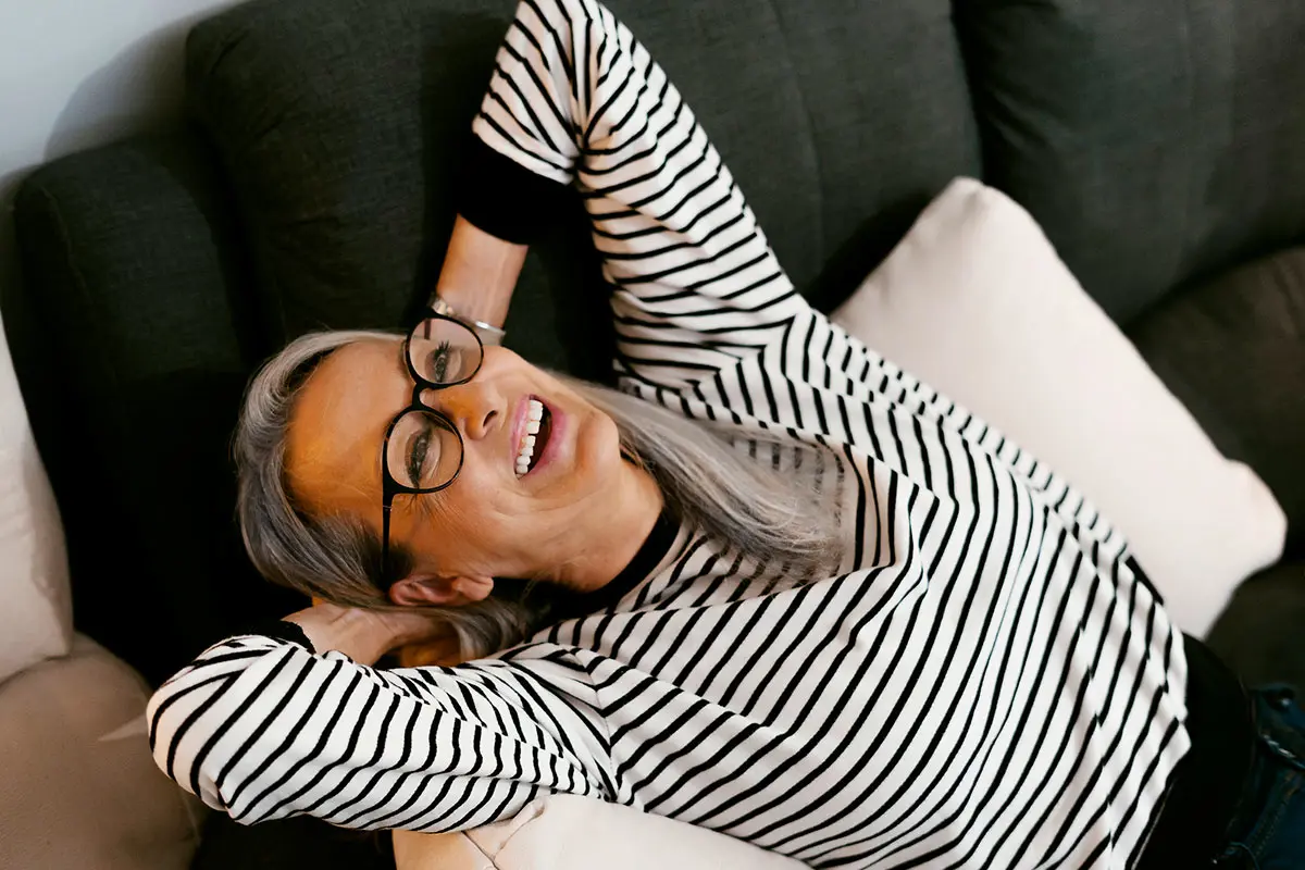 Woman with long grey hair in glasses and striped shirt leaning back in happiness on sofa. AW296