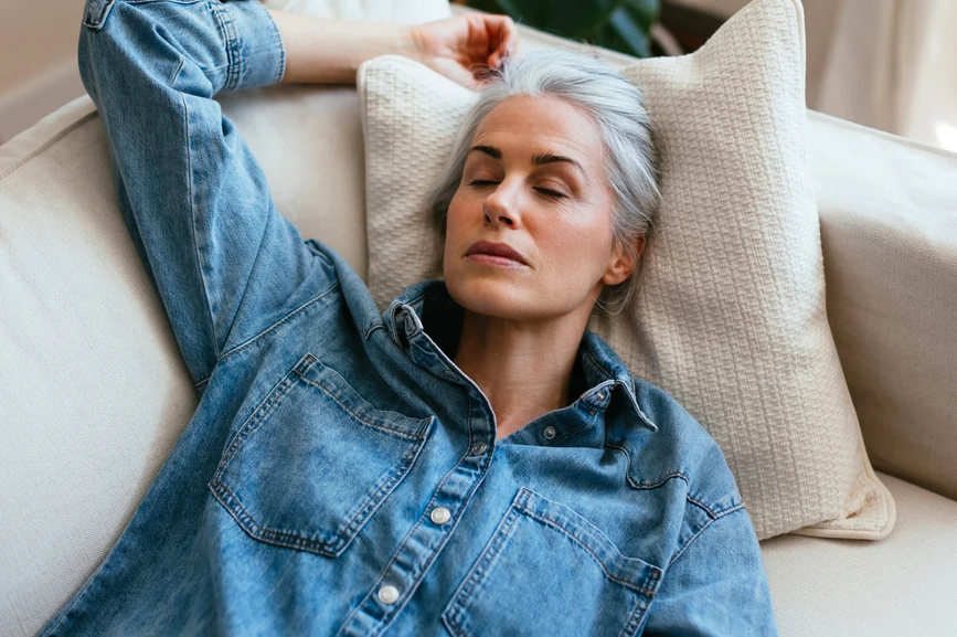 Grey haired youthful looking woman resting on sofa in denim shirt, eyes closed. AW071