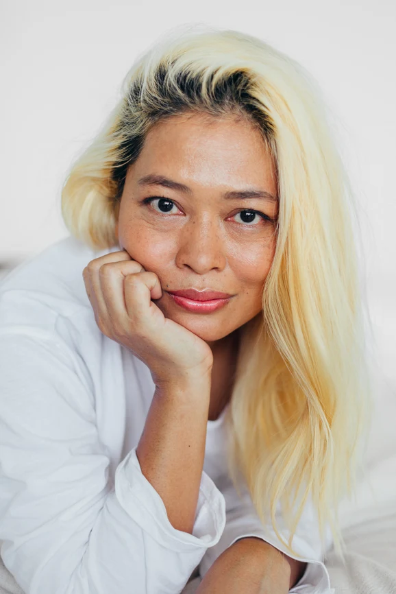 Asian woman with bleached blonde hair resting her head on hand supported by elbow in white shirt. AW228