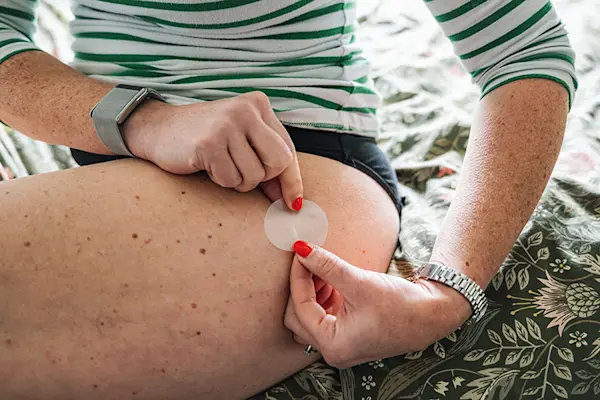Detail of a woman in a striped shirt, sitting in the edge of her bed, applying transdermal patch to her thigh. thumbnail