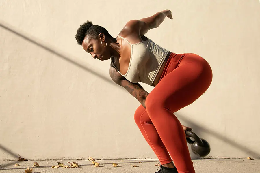 A Woman in orange leggings and white tank top is demonstrating a twist using a kettlebell in one hand while in a bent legged position. (inline)