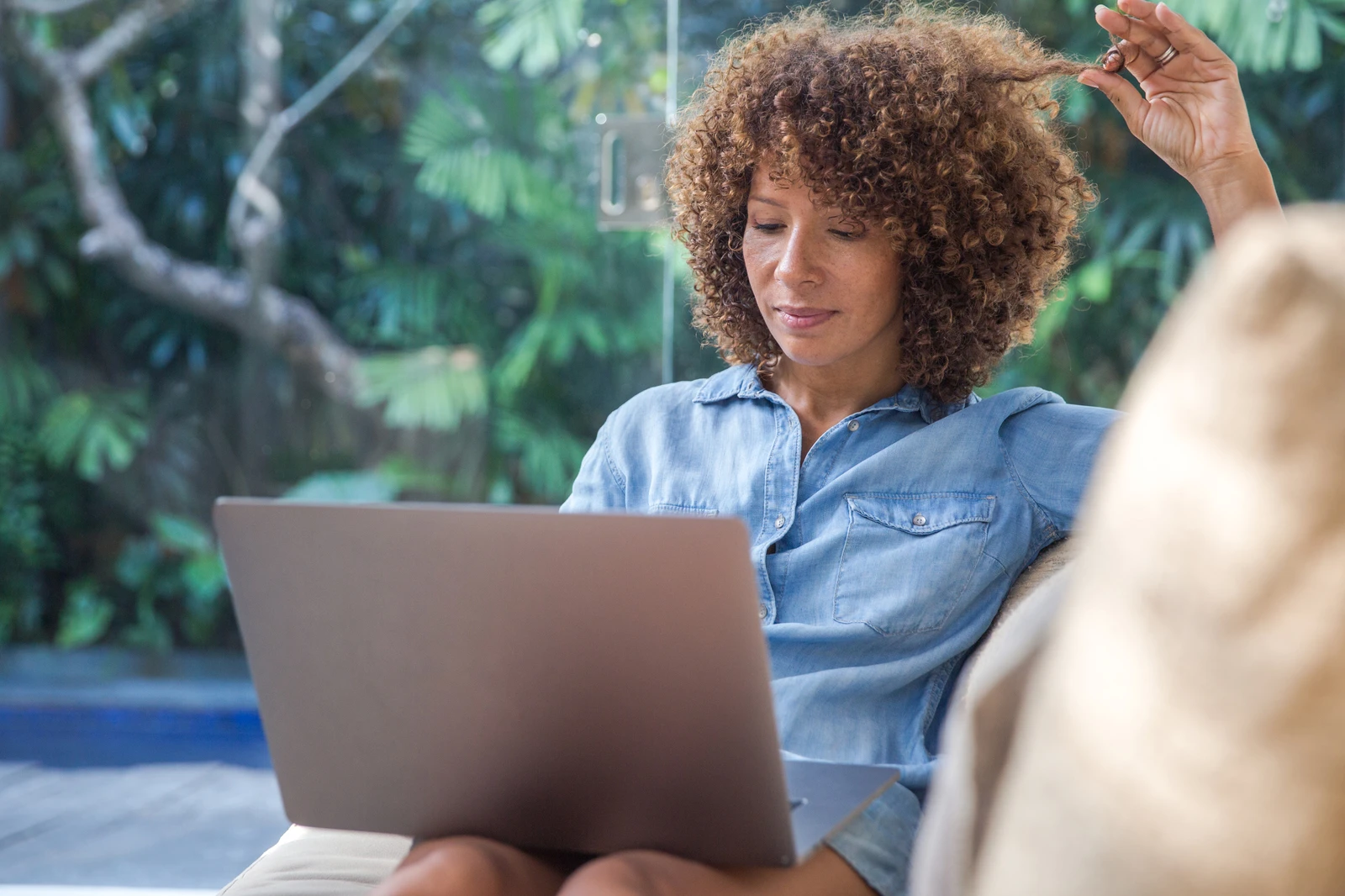 Bi-racial woman looking at laptop on sofa absentmindedly playing with curls. AW086