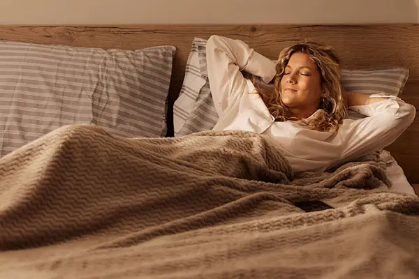 Woman resting comfortably in bed at night with eyes closed, arms behind her head.