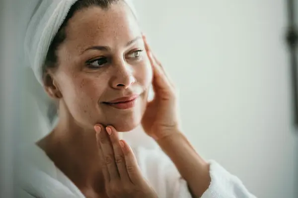 Woman with towel wrapped around her hair looking in the mirror touching her face.  AW67 thumbnail