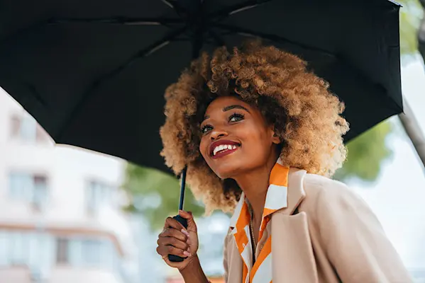 Smiling black woman with golden blonde hair carrying umbrella, looking into distance. thumbnail