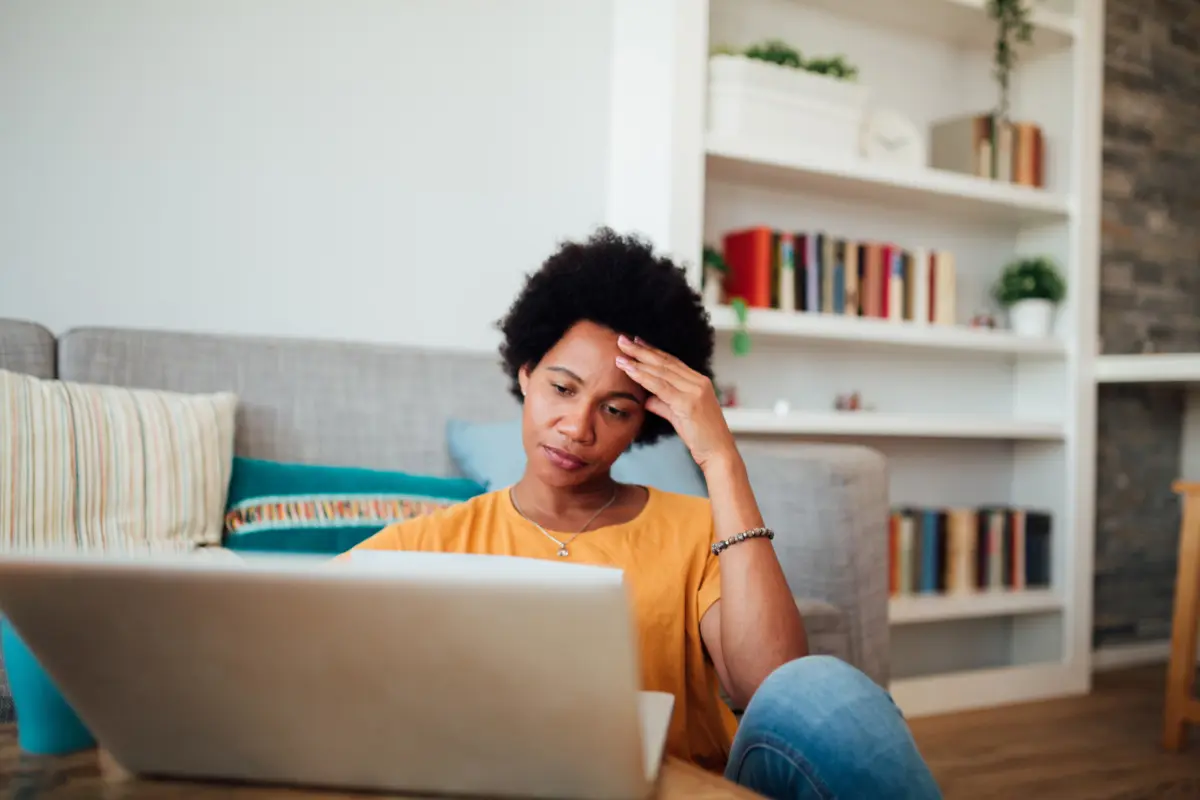 Black woman sitting on the floor at laptop with hand to forehead in discomfort. AW502 