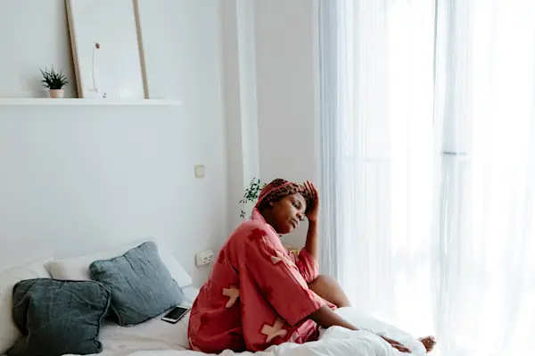 Woman of color in robe sitting on bed in morning light filtered through sheer curtains. AW117