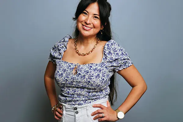 Hispanic woman with her hands on her hips in floral top in front of blue studio backdrop.  thumbnail