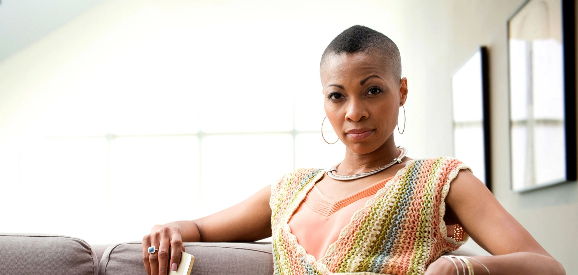 Black woman with short hair on sofa wearing open knit striped sweater vest, looking at camera with serious expression. AW247