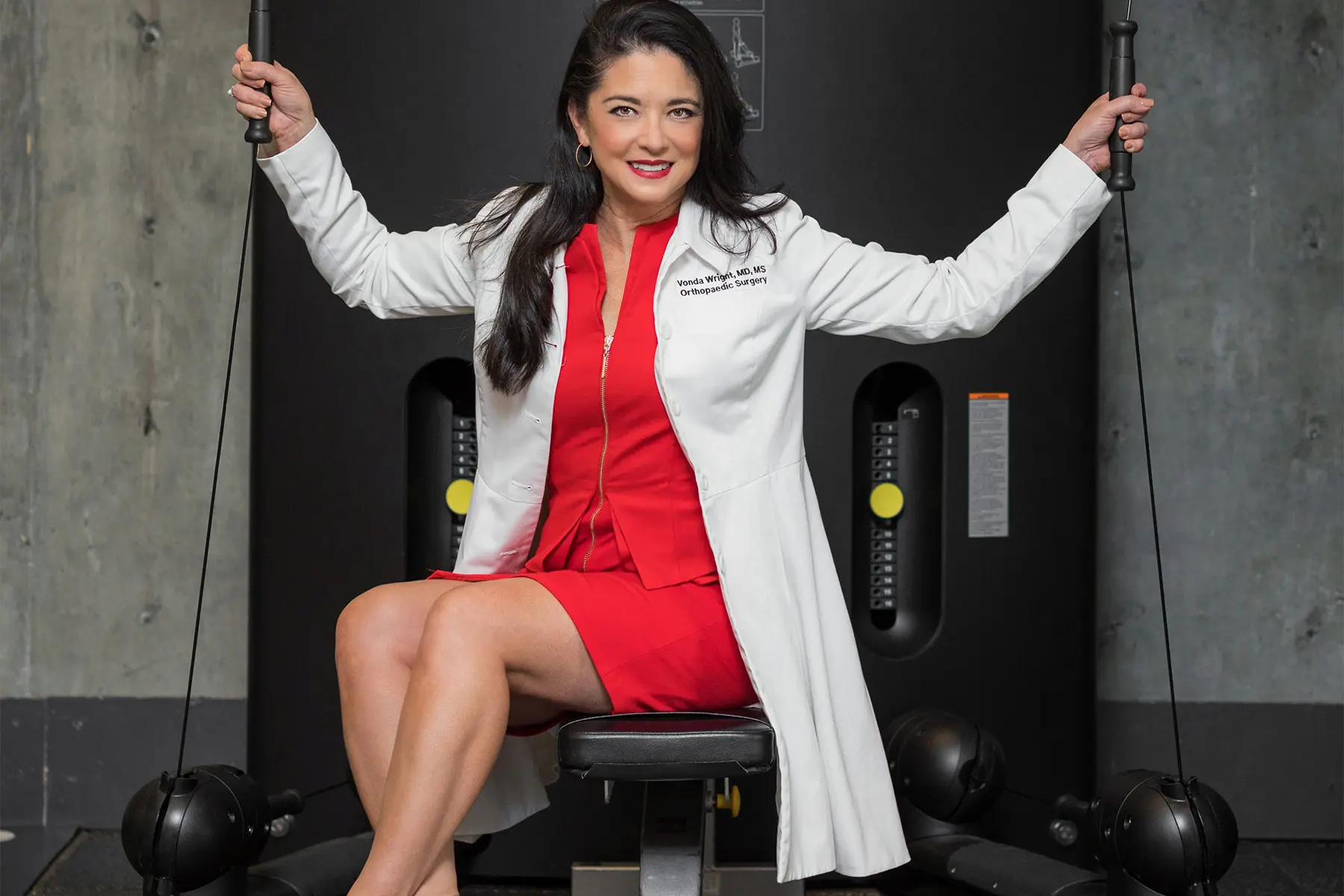 Dr. Vonda Wright posing on exercise equipment wearing lab coat over a red suit.