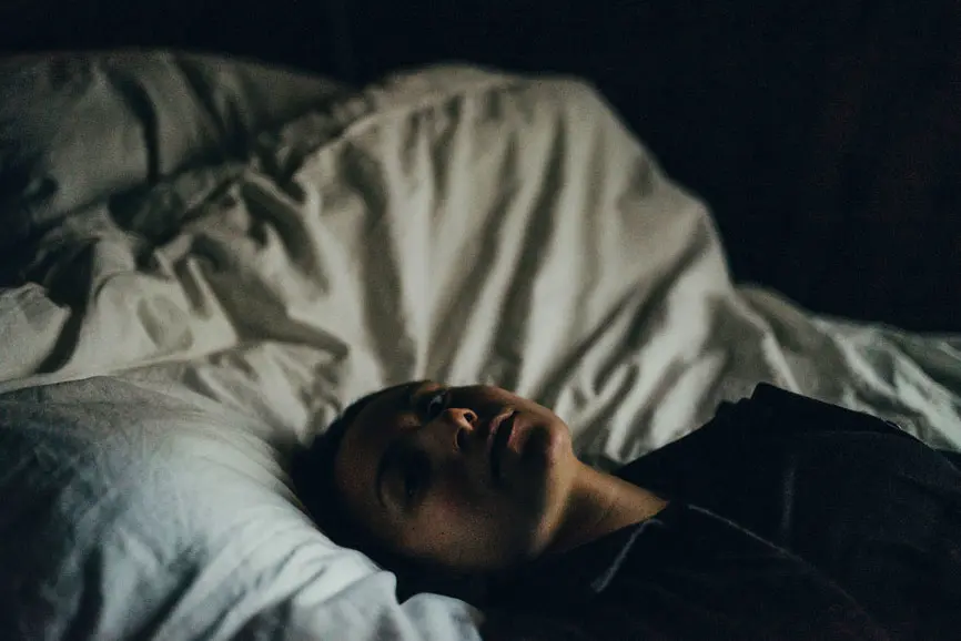 Woman in darkened room on bed, eyes open. AW191 