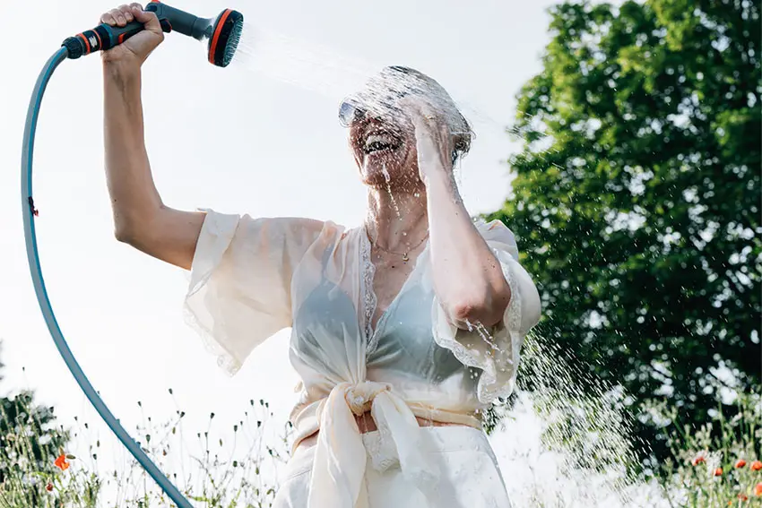 Woman in sunglasses and white blouse, cooling down with spray from garden hose, smiling. AW222 