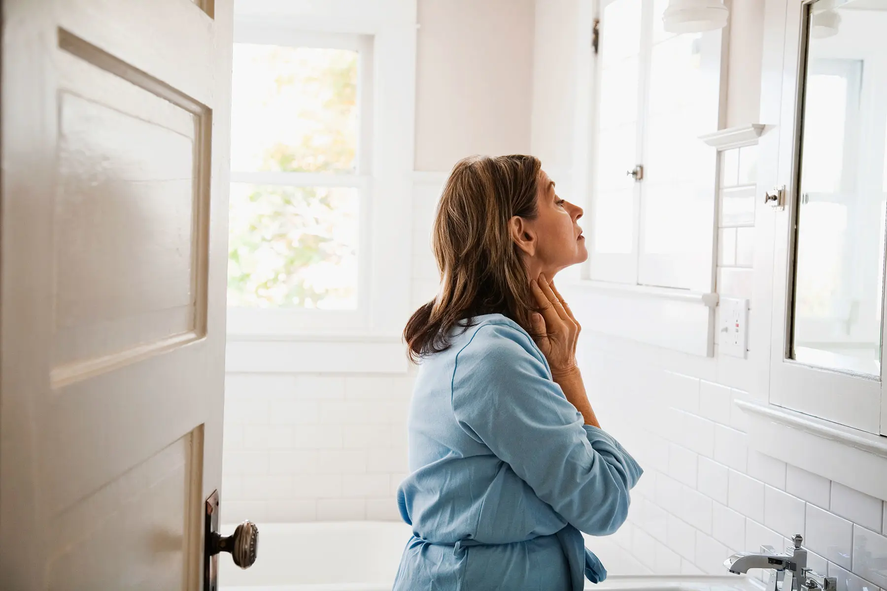 Woman in bathrobe looking in bathroom mirror applying cream to neck. AW548