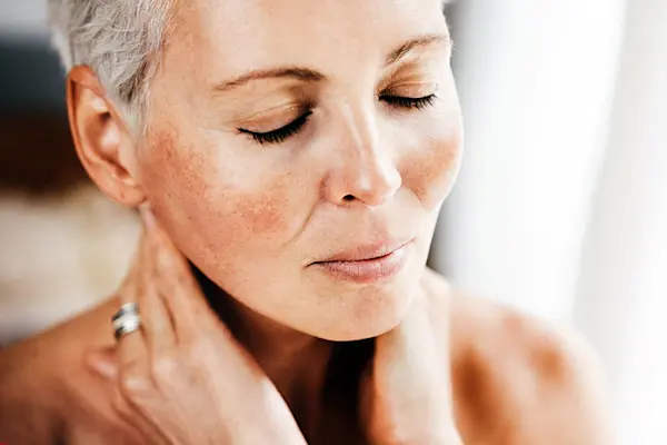Middle aged woman with rosacea on her cheeks, with eyes closed and her hands touching her neck. thumbnail