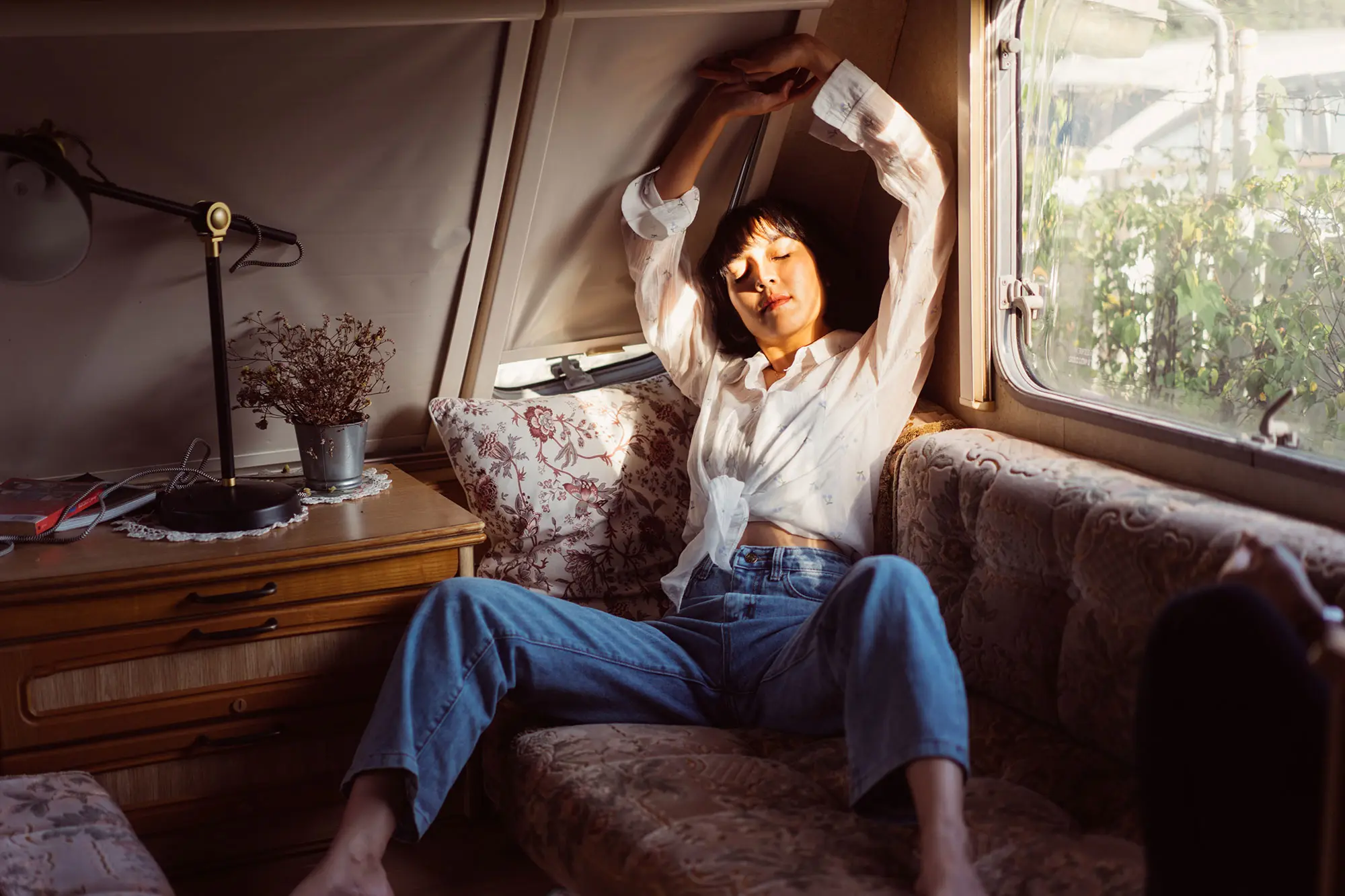 Woman bathed in light in a vintage camper wearing jeans, stretching with closed eyes. AW380