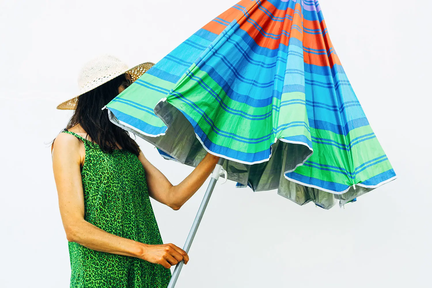 Woman in a green sleeveless dress and straw hat opening colorful beach umbrella. AW525