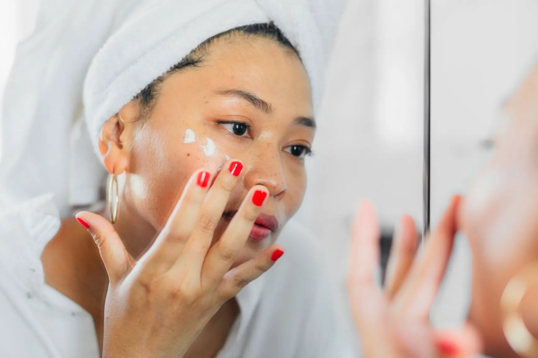 Asian woman applying dots of cream to cheek while looking in the mirror, wearing towel around her hair. AW450