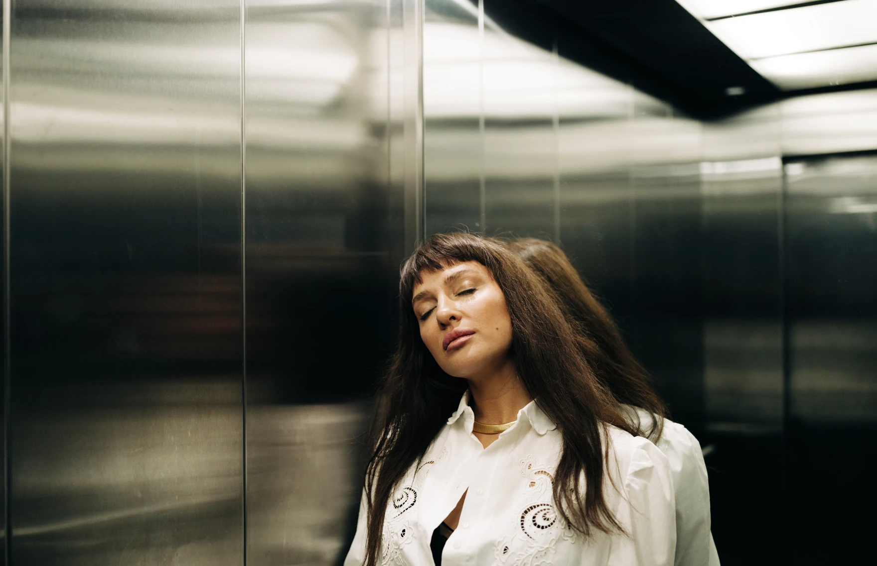 Woman lwith long brunette hair and bangs leaning on wall inside an elevator, eyes closed. AW183