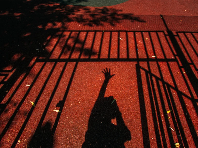 Woman's shadow on red pavement, arm reaching up in anguish with shadow of gate around her.  AW184 