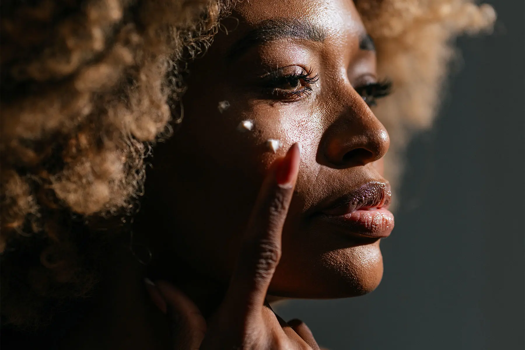 Mature Black woman with gold curls applying dots of cream to her face. AW615