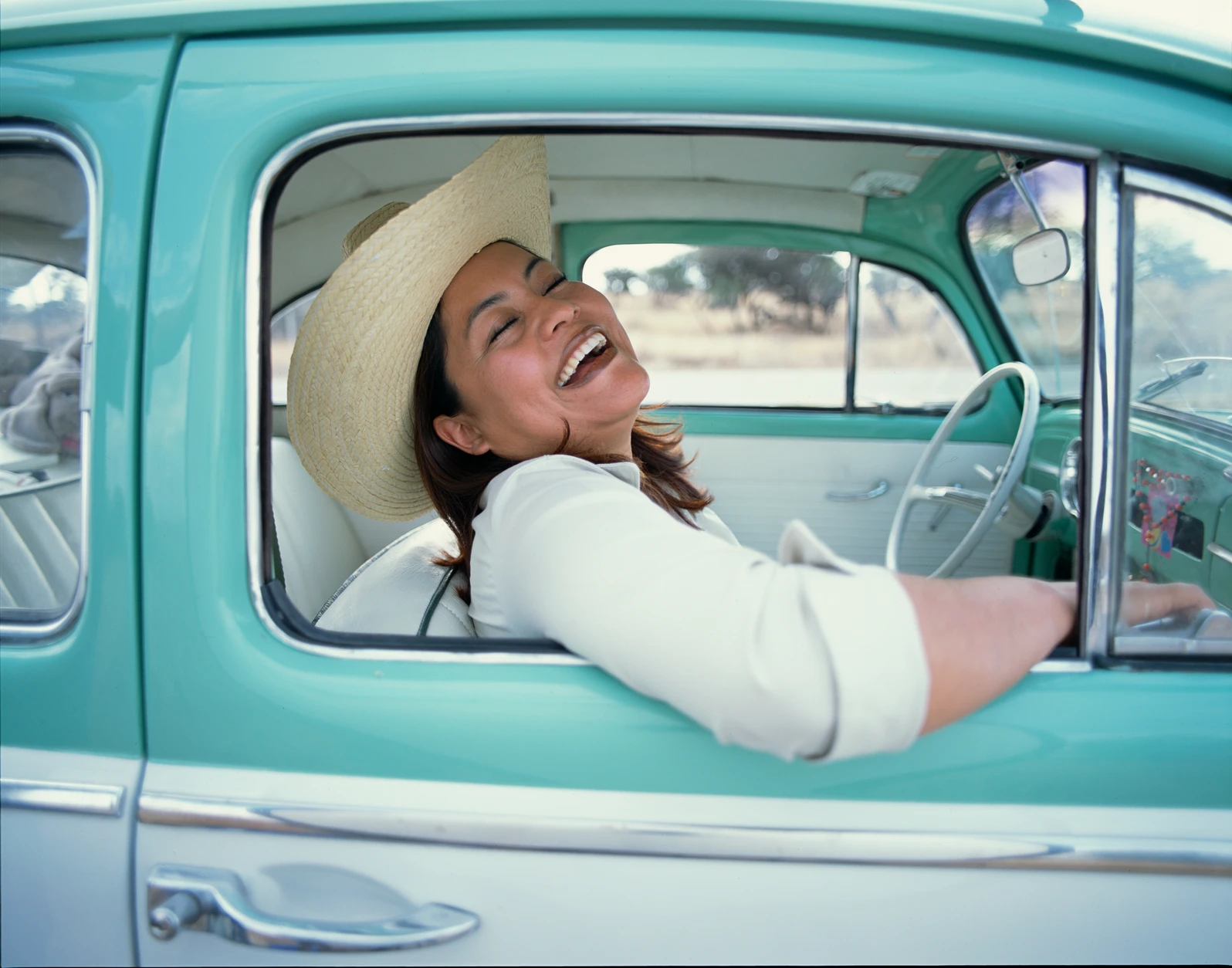 Latina woman wearing cowboy hat sitting in vintage car, laughing. AW341 