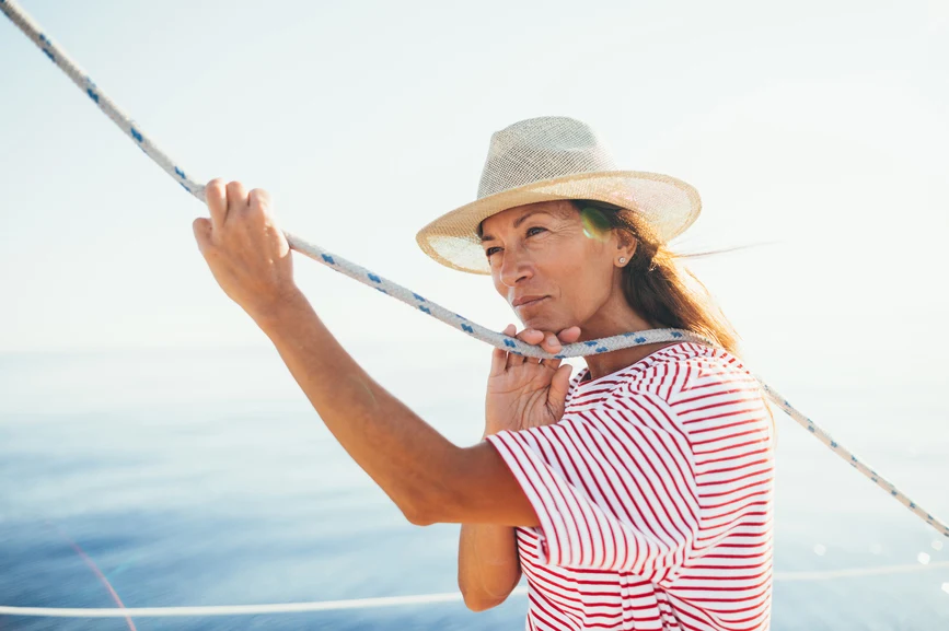 Woman in hat and striped shirt on a boat at sea holding a sail rope in thought. AW139