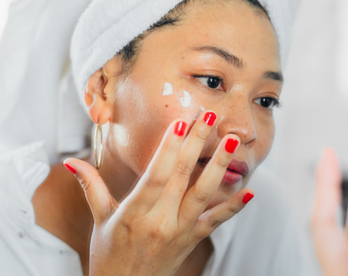 Asian woman with red nail polish, hair wrapped in towel applying dots of cream to her face. AW450