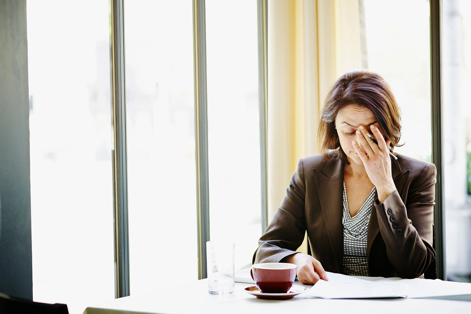 Woman wearing blazer sitting at desk with cup of coffee rubbing her forehead. AW483