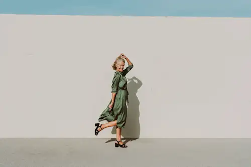 Woman in her mid to late 50s in chunky high heels and green satin dress walking playfully in front of stucco wall. header