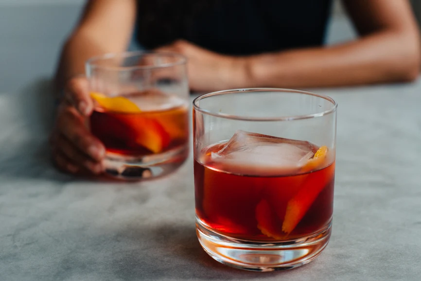 Two cocktails on tabletop with woman out of focus holding one glass.  AW200